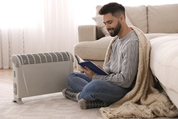 Young man warming up near electric heater at home