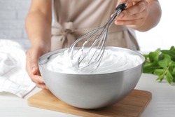 Woman whipping white cream with balloon whisk at wooden table, closeup
