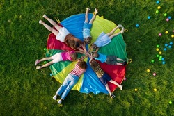Group of children with teachers holding hands together on rainbow playground parachute in park, top view. Summer camp activity