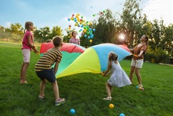 Group of children and teachers playing with rainbow playground parachute on green grass. Summer camp activity