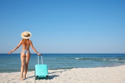Woman in bikini with suitcase on sandy beach near sea, back view