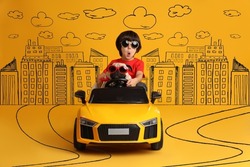 Cute little boy with his dog in toy car and drawing of city on yellow background