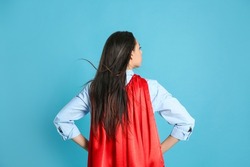 Young woman wearing superhero cape on light blue background, back view