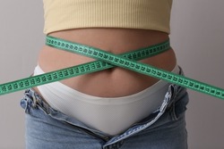 Woman in unfit jeans with measuring tape on light grey background, closeup