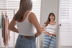 Young woman trying to put on tight jeans near mirror at home