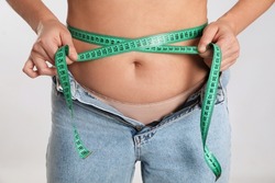 Woman in unfit jeans measuring her waist on light background, closeup. Weight loss concept