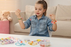 Cute little girl making beaded jewelry at table in room