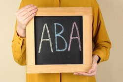 Woman holding small chalkboard with abbreviation ABA (Applied behavior analysis) on beige background, closeup