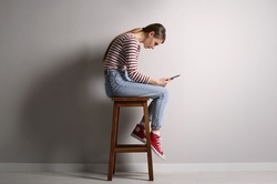 Woman with bad posture using tablet while sitting on stool near light grey wall indoors