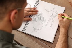 Man drawing in sketchbook with pencil at wooden table, closeup