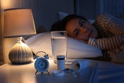 Mature woman suffering from insomnia in bed at night, focus on nightstand with pills, alarm clock and water