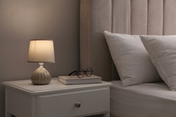 Stylish lamp, book and glasses on bedside table indoors. Bedroom interior elements