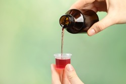 Woman pouring cough syrup into measuring cup on light green background, closeup