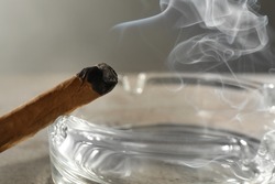 Smoldering cigar near glass ashtray on light grey table, closeup