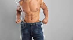 Closeup view of man with muscular body in oversized jeans on grey background, space for text. Weight loss