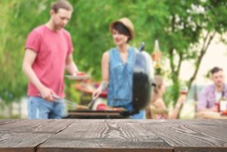 Empty wooden table and blurred view of people having barbecue with modern grill outdoors