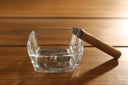 Smoldering cigar near glass ashtray on wooden table, closeup