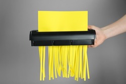 Woman destroying sheet of yellow paper with shredder on grey background, closeup