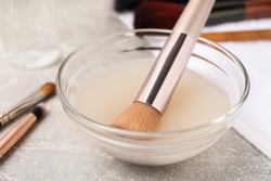 Makeup brush in bowl with cleanser on grey table, closeup