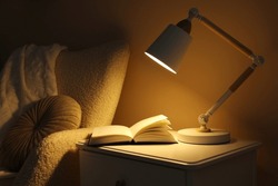 Stylish modern desk lamp and open book on white cabinet near armchair in dark room