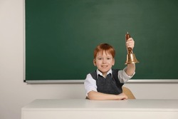 Cute little boy ringing school bell in classroom, space for text