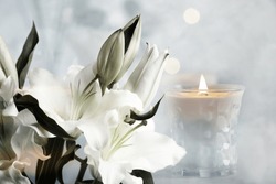 Funeral. Beautiful lilies and burning candle on light blurred background, bokeh effect.