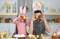 Father and his cute daughter covering eyes with beautifully painted Easter eggs at table in kitchen