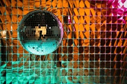 Shiny disco ball against foil party curtain under turquoise and orange light, space for text