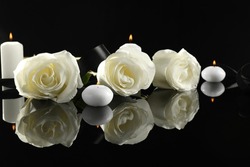 White roses and burning candles on black mirror surface in darkness. Funeral symbols