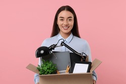 Happy unemployed woman holding box of personal office belongings on pink background
