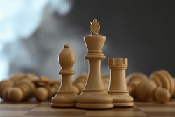 Wooden king, rook and bishop in front of fallen chess pieces on checkerboard against dark background, closeup