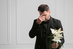 Sad man with lily flowers mourning near white wall, space for text. Funeral ceremony