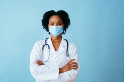 Portrait of a doctor in lab coat with face mask and arms crossed, isolated on blue background