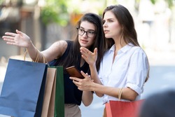 A woman carrying a shopping bag telling Give directions in the city for a woman hold a mobile phone. Young beautiful woman walking after successful shopping.