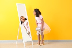 Adorable little African-American girl with mother's shoes looking into mirror near color wall