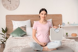 Young pregnant woman with measuring scales in bedroom
