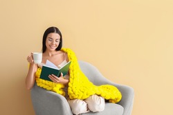 Beautiful young woman with warm plaid drinking tea and reading book while sitting in armchair on color background