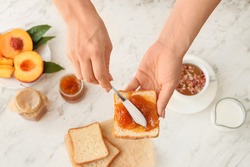 Woman spreading tasty peach jam onto slice of bread on light background