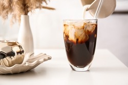 Pouring milk from jug into glass with tasty ice coffee on table, closeup