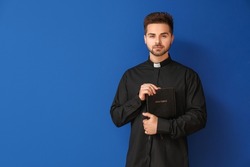 Young priest with Bible on color background
