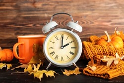 Alarm clock and autumn leaves with cup of tea and clothes on wooden background. Daylight saving time end