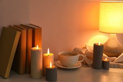 Burning candles, cup of coffee, books and glowing lamp on table near wall