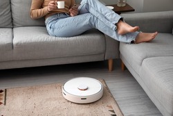 Modern robot vacuum cleaner near sofa with resting woman in room