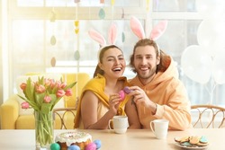 Happy couple in bunny ears cracking Easter eggs at table