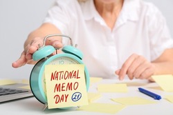 Alarm clock and sticky note paper with text NATIONAL MEMO DAY on table of mature secretary