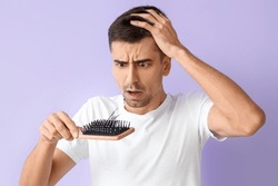 Worried young man holding brush with fallen down hair on lilac background