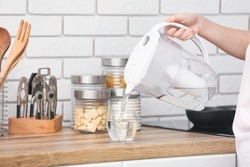 Woman pouring purified water into glass from filter jug in kitchen