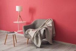 Comfortable armchair with plaid, tables and lamp near pink wall in room