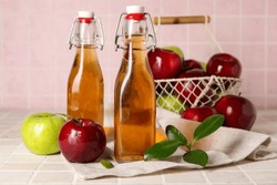 Glass bottles of fresh apple cider vinegar and basket with fruits on pink tile table