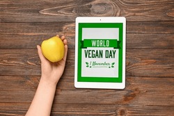 Female hand with apple and digital tablet with text WORLD VEGAN DAY on wooden background